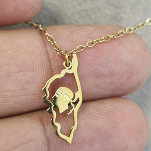 Puede incluir: Un collar de tono dorado con un colgante en forma de la isla de Córcega. El colgante es un contorno de metal fino que contiene una silueta recortada de la cabeza de moro, un símbolo tradicional, suspendido de una delicada cadena dorada.