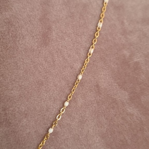 Peut inclure: Un collier en cha&icirc;ne dor&eacute;e avec un petit pendentif en forme de croix. La cha&icirc;ne est parsem&eacute;e de petites perles blanches.
