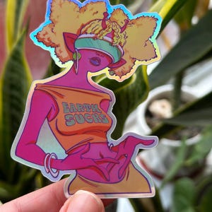Op de afbeelding: Een holografische sticker met een roze cartoonfiguur met geel haar, die een futuristische vizier draagt en een geel shirt met de tekst "Earth Sucks".