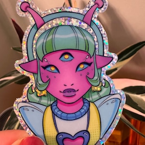 Puede incluir: Una pegatina holográfica con un extraterrestre rosa con cabello verde, tres ojos y un colgante en forma de corazón. El extraterrestre lleva un traje espacial azul y amarillo.