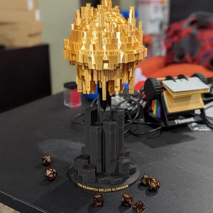 May include: A gold-colored, 3D-printed sculpture with a detailed, geometric design. The sculpture sits atop a black base with the text "PAX MAGNIFICA BELLUM GLORIOSUM". Several dice are scattered around the base, suggesting a tabletop game theme.