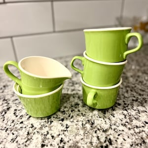 Peut inclure: Ensemble de tasses et d'une crémière en céramique verte, avec des rebords et des anses blancs. Les tasses sont empilées, et la crémière est à côté. L'ensemble est sur un comptoir moucheté.