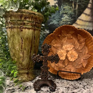 Könnte beinhalten: Eine dekorative Holzschale mit einem geschnitzten Blumenmuster steht auf einem schwarzen Ständer. Ein kleines, verziertes Metallkreuz steht vor der Schale. Eine grüne Keramikvase mit braunen Streifen befindet sich im Hintergrund.