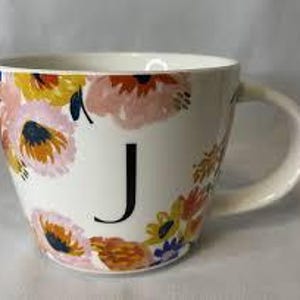 Puede incluir: Taza de cerámica blanca con un diseño floral en tonos rosas, amarillos y naranjas. Una gran letra negra "J" está centrada en la taza. La taza tiene un asa curva y una abertura ancha.