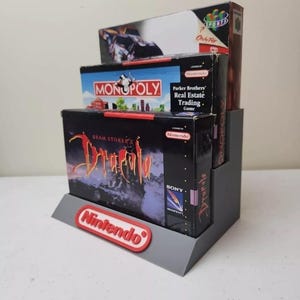 May include: A gray plastic display stand with three video game cartridges. The top cartridge is for the Nintendo game "Only For Sports". The middle cartridge is for the Nintendo game "Parker Brothers' Real Estate Trading Game". The bottom cartridge is for the Nintendo game "Bram Stoker's Dracula".