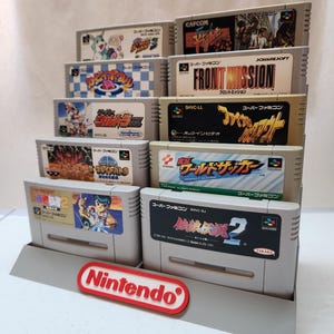 May include: A gray tiered display stand holding several Super Famicom video game cartridges. The cartridges feature colorful labels with Japanese text and graphics. A red and white Nintendo logo is at the base of the stand.