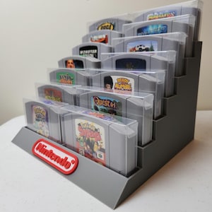 May include: A gray plastic display stand for Nintendo 64 game cartridges. The stand has five tiers and holds 15 cartridges. The cartridges are in clear plastic cases and feature various game titles, including "Ready Rumble", "Quest 64", and "Pokemon Snap". The stand has a red "Nintendo" logo on the front.