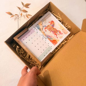 May include: A 2026 calendar with a wooden frame, featuring a watercolor illustration of a horse. The calendar is inside a cardboard box with paper filler. The month of April is visible.