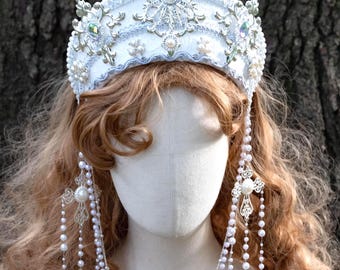 Op de afbeelding: Een witte en zilveren bruidskroon met parel- en kristalaccenten. De kroon heeft sierlijke details en wordt gecombineerd met bijpassende parel- en kristallen oorbellen en een gelaagde ketting. Het hoofddeksel wordt getoond op een mannequinhoofd.