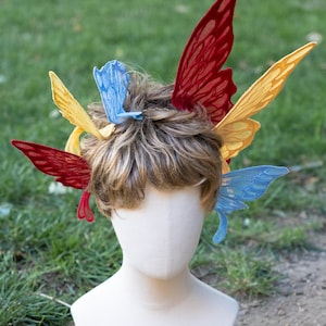 May include: A white mannequin head with short brown hair adorned with multiple fabric butterfly wings in vibrant shades of red, yellow, and blue. The wings are arranged around the hair, featuring intricate vein patterns and translucent textures.