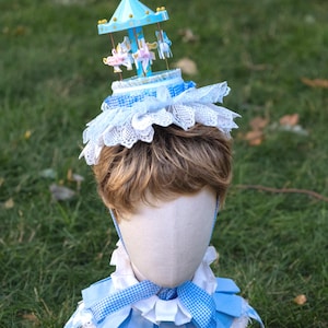 May include: A headpiece featuring a miniature blue carousel with white horses, adorned with white lace and blue gingham fabric. Below, a matching blue and white pleated collar with gingham accents and white pom-poms completes the set.