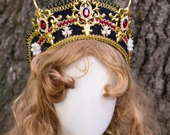 May include: A black fabric crown featuring ornate gold metal filigree, red oval-shaped gemstones, and clusters of small white pearls. The crown is adorned with intricate gold braided trim and sits atop a mannequin head with wavy blonde hair.