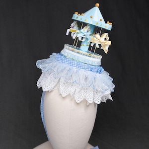 May include: A whimsical hat featuring a miniature carousel design. The hat has a light blue conical top with gold accents and a carousel with white horses. It is adorned with a white lace and blue gingham trim, and a matching gingham ribbon.