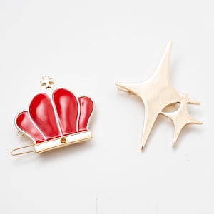 May include: Two gold hair clips, one shaped like a crown with red enamel and a single jewel, the other shaped like a double star.
