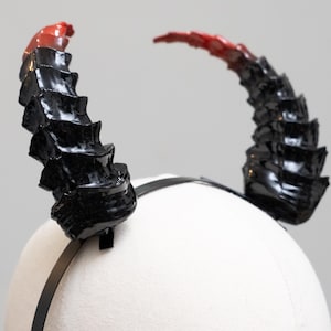 May include: A pair of black and red devil horns attached to a black headband. The horns are glossy and have a textured, ridged design. The tips of the horns fade from black to red.
