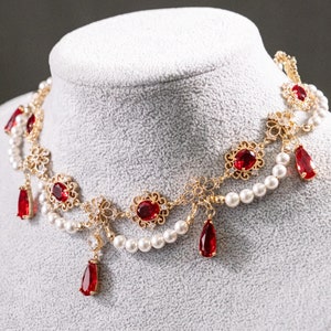 May include: A gold necklace with red gemstone accents and white pearls. The necklace has a delicate, intricate design and features a dangling drop pendant.