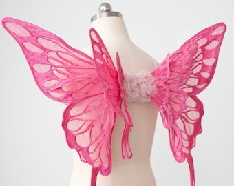 May include: Pink butterfly wings made of sheer fabric with a delicate lace design. The wings are attached to a white dress form.