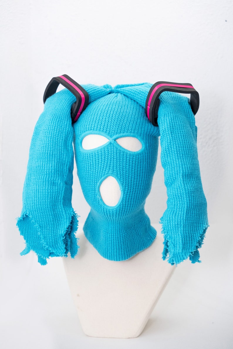 May include: A blue knitted balaclava with two long blue knitted strands attached to the top, resembling pigtails. The balaclava has two eye holes and a mouth hole. The pigtails have black and pink bands around them.