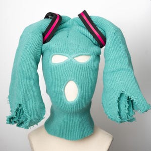 May include: A teal blue ski mask with two long teal blue tails, resembling bunny ears, and black and pink elastic bands on top.