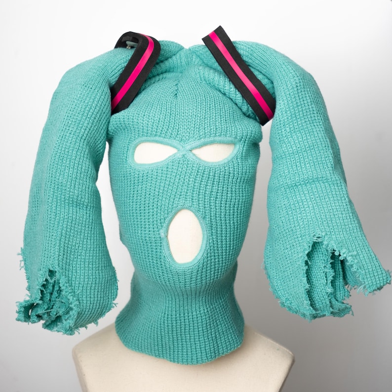 May include: A teal blue ski mask with two long teal blue tails, resembling bunny ears, and black and pink elastic bands on top.