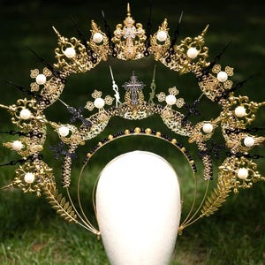 May include: An ornate gold and black headpiece with crosses, pearls, and intricate filigree designs. The crown-like structure features a halo shape with multiple points and a central cross. The headpiece is displayed on a white mannequin head against a green background.