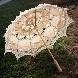 May include: An open, cream-colored lace parasol with a wooden handle rests on grass. The parasol features intricate floral and scalloped lace patterns. The wooden handle is light brown with a carved grip.