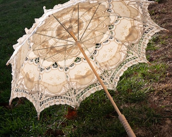 May include: An open, cream-colored lace parasol with a wooden handle rests on grass. The parasol features intricate floral and scalloped lace patterns. The wooden handle is light brown with a carved grip.