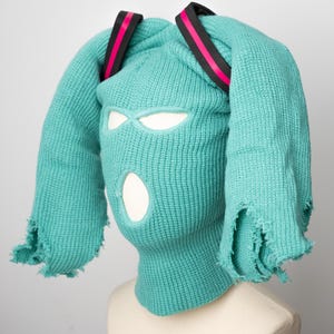 May include: A turquoise knitted balaclava with two long, frayed strands of fabric extending from the top. The balaclava has eye and mouth holes and is accented with two black and pink elastic bands.