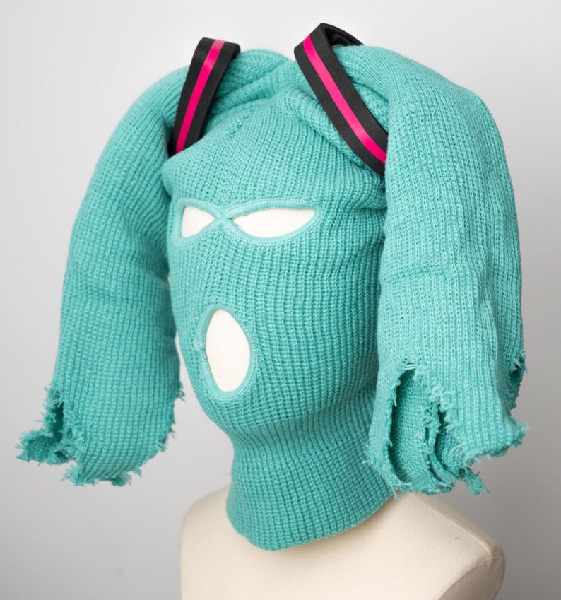 May include: A turquoise knitted balaclava with two long, frayed strands of fabric extending from the top. The balaclava has eye and mouth holes and is accented with two black and pink elastic bands.