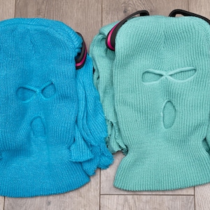 May include: Two knitted balaclavas, one blue and one green, with eye and mouth holes. Both have a black band around the top.