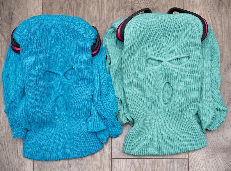 May include: Two knitted balaclavas, one blue and one green, with eye and mouth holes. Both have a black band around the top.