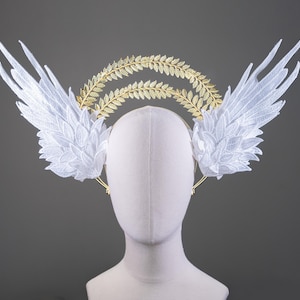 May include: A white lace angel wing headband with a gold laurel wreath. The headband is on a white mannequin head.