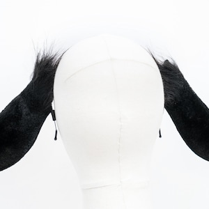 May include: Black furry animal ears headband. The headband is made of black faux fur and has two long, floppy ears. The ears are attached to a black elastic headband.