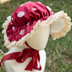 May include: A red bonnet with white polka dots and a lace trim. The bonnet has a red ribbon tied at the back.