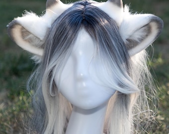May include: A pair of white and gray faux fur animal ears with a matching wig. The ears are detailed with gray tips and inner ear accents. The wig features long, layered, gray and white hair, styled to frame the face.