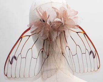 May include: A headpiece with translucent, veined wings and a cluster of pink fabric flowers. The wings are outlined in brown and have a reddish-brown border. The headpiece is mounted on a white mannequin head.