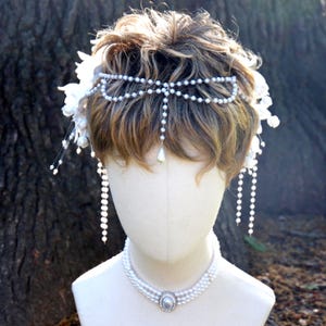 May include: A bridal headpiece and necklace set. The headpiece features a delicate pearl design with dangling strands. The necklace is a multi-strand pearl design with a central oval pendant. White floral accents are also included.