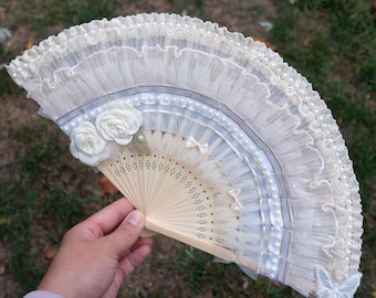 May include: A cream-colored hand fan with a delicate design. The fan features layers of lace, pearls, and small white bows. Two white roses and a butterfly accent the fan's design. The fan is made of wood and fabric.