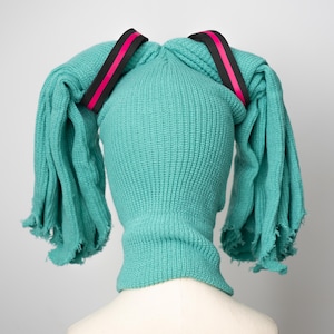 May include: A teal blue knitted hat with two long teal blue tails tied together with black and pink ribbon.