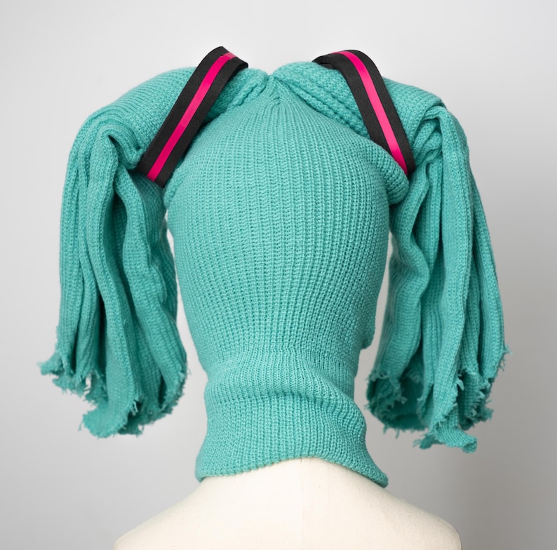 May include: A teal blue knitted hat with two long teal blue tails tied together with black and pink ribbon.