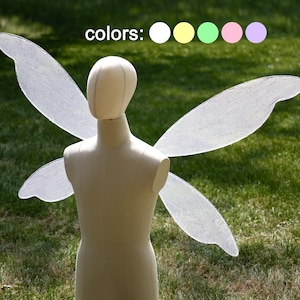May include: A mannequin wearing a pair of clear, glittery fairy wings. The wings are attached to the mannequin's back and are spread out. The text "colors:" is above a series of colored circles: white, yellow, green, pink, and purple.