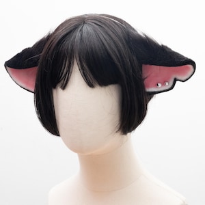 May include: Black cat ear headband with pink inner ears and silver stud accents. The ears are fluffy and attached to a black wig. The wig has short, straight black hair.
