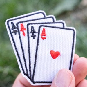 May include: An embroidered patch featuring four white playing cards arranged in a fan shape. Each card displays a black spade or red heart suit symbol and a corresponding letter A. The design is outlined in black thread on a white fabric base.