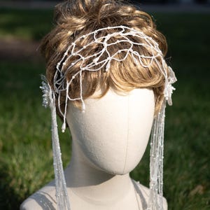 May include: A bridal headpiece on a mannequin head. The headpiece is white and features a delicate, web-like design across the top, with strands of beads cascading down. The hair is styled in a light brown updo.