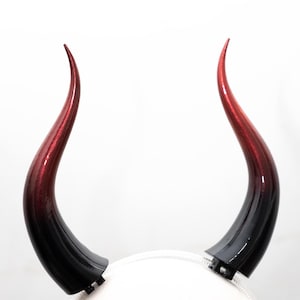 May include: A pair of devil horns on a white headband. The horns are curved, with a gradient of red to black. The horns are glossy and appear to be made of plastic.