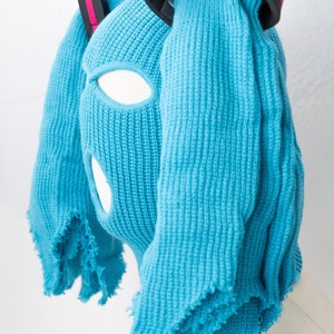May include: A blue knitted balaclava with two long, fringed strands of fabric attached to the top. The balaclava has eye and mouth holes. The strands of fabric are held together by a black headband with a pink stripe.