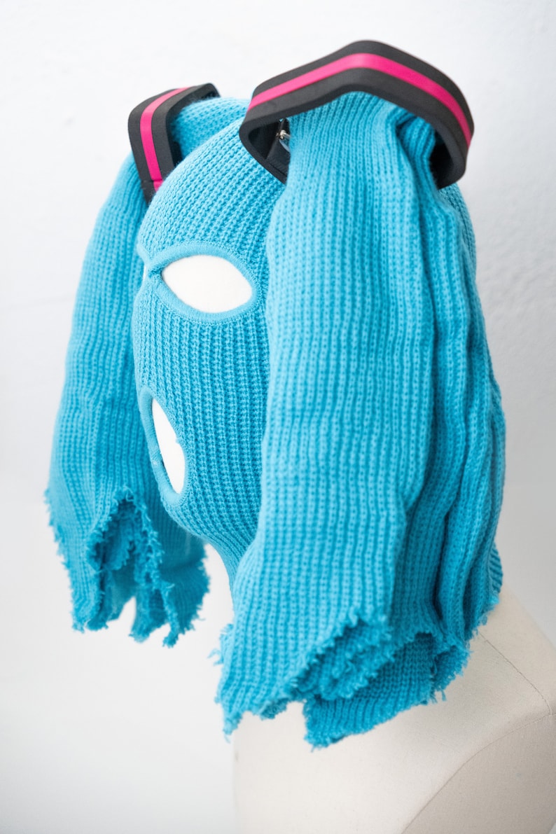 May include: A blue knitted balaclava with two long, fringed strands of fabric attached to the top. The balaclava has eye and mouth holes. The strands of fabric are held together by a black headband with a pink stripe.