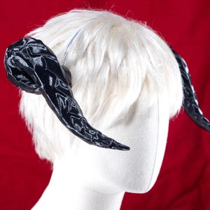 May include: A pair of black, curved horns with a textured pattern, attached to a white wig. The horns are positioned on a mannequin head against a red background. The horns are a costume accessory.