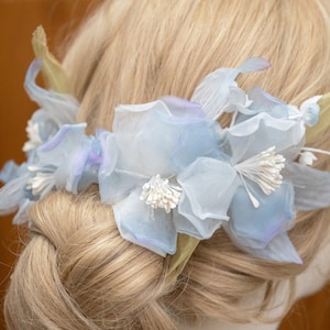 May include: A light blue flower hair comb with white accents. The comb is made of fabric and is designed to look like a flower.