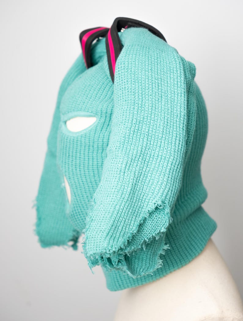 May include: A turquoise knitted balaclava with a frayed edge and a black strap with pink accents.